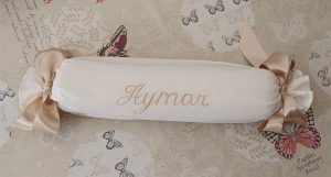 Cojín artesanal blanco en forma de caramelo, con nombre bordado y lazos beige